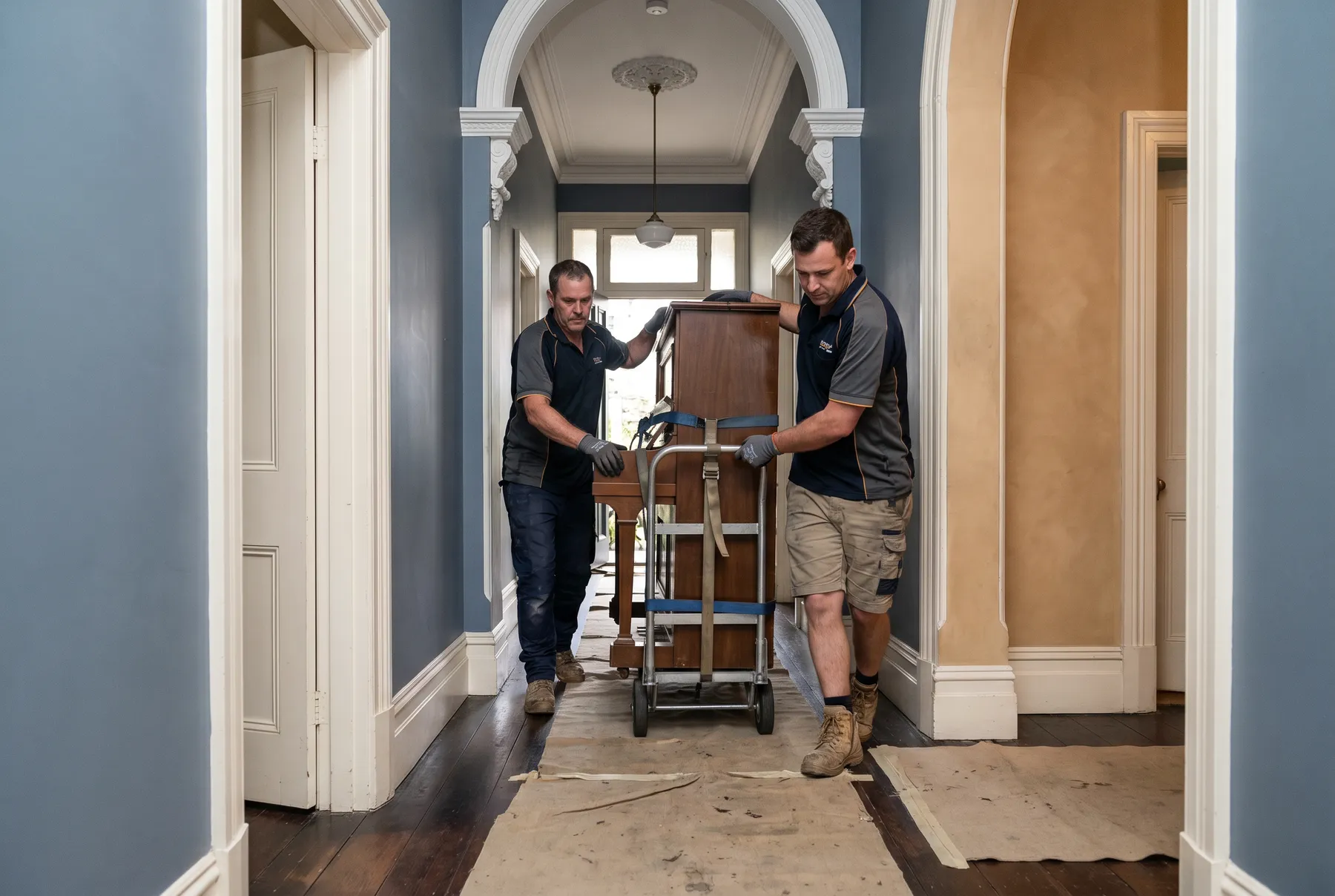 Two removalists easing an upright piano along a period hallway